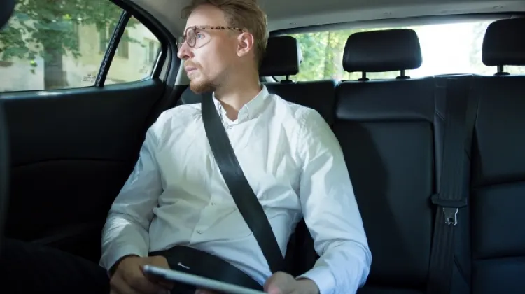 車の後部座席でシートベルトを締め、窓の外を見つめる男性。手にはタブレットを持ち、移動中の様子を表している。