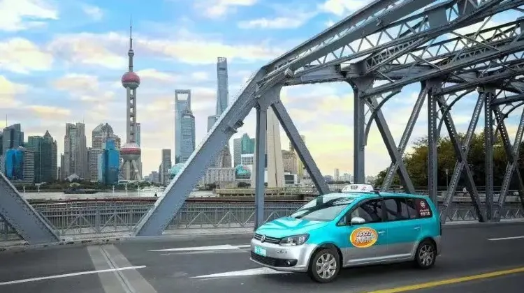 外灘エリアの橋を走る青色の上海タクシー。背景に上海タワーや東方明珠タワーが見える市街地の風景。