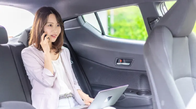 車の後部座席でノートパソコンを開き、電話をかけながら仕事をする女性。移動中のビジネスシーンを表している。